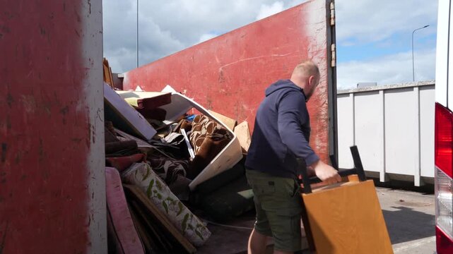 Man in hoodie and shorts hurls and stacks old furniture units into a red metal skip at a public dump site.