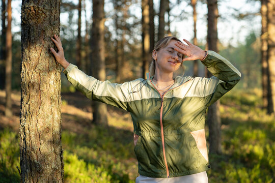 Joyful woman hiker enjoying forest walk and gaining positive emotions, standing beside tree smiling, cheerful relaxed female stretching body while resting in woodlands, peaceful moment in nature.