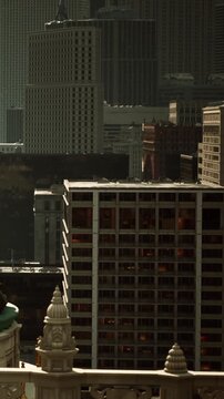 The city skyline displays a mix of modern and classic buildings. Sunlight reflects off glass windows as people move below. The scene captures daily life in a busy urban area.