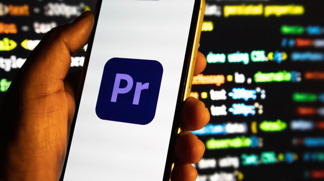 Closeup of a hand holding a smartphone displaying the adobe premiere pro logo against a blurred background of computer code