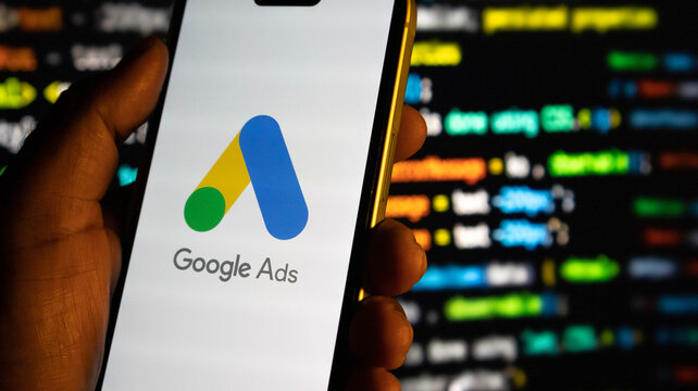 Closeup of a hand holding a smartphone displaying the google ads logo against a background of computer code