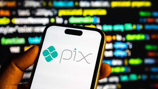 Closeup of a hand holding a smartphone displaying the pix payment system logo against a blurred background of computer code