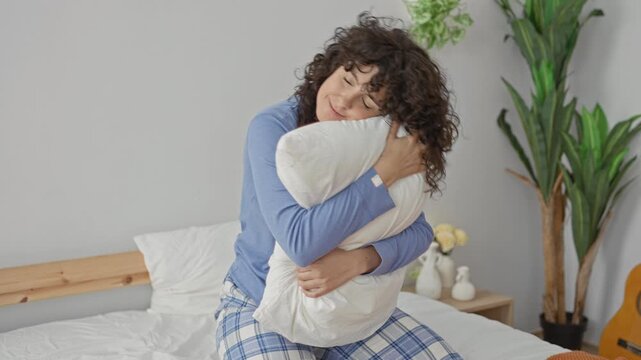 Woman hugging a white pillow, eyes closed, hands clutching cushion, sitting on bed in a bedroom with plant and guitar visible; comfort.