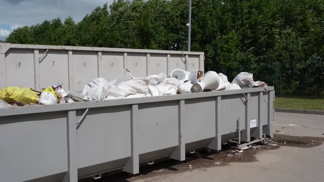 Gray metal skip overflows with white rubble sacks, broken toilets, and mixed junk at an outdoor civic amenity yard.