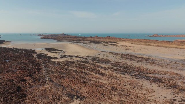 Cobo Bay Guernsey Channel Islands.Panning view over rugged shoreline of golden sandy beach and jetty out to sea over pink granite rocks towards calm sea and rocky outcrops on sunny day with blue sky
