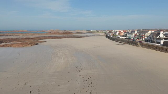 Cobo Bay Guernsey Channel Islands.Forward drone footage over extensive golden sandy beach and seafront buildings with views across rocky outcrops out to sea and horizon on bright sunny day