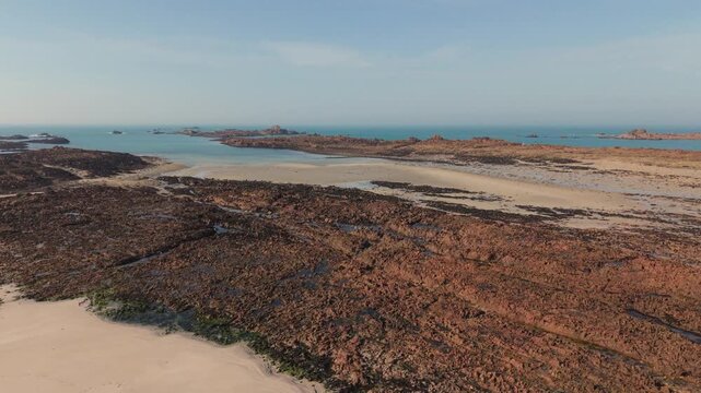 Cobo Bay Guernsey Channel Islands.Low panning view over rugged shoreline of golden sandy beach out to sea over pink granite rocks towards calm sea and rocky outcrops on sunny day with blue sky