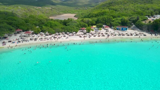 Aerial parallax revealing stunning Grote Knip beach in Curacao with vivid turquoise bay, white sand, crowds of swimmers and lush green hillsides