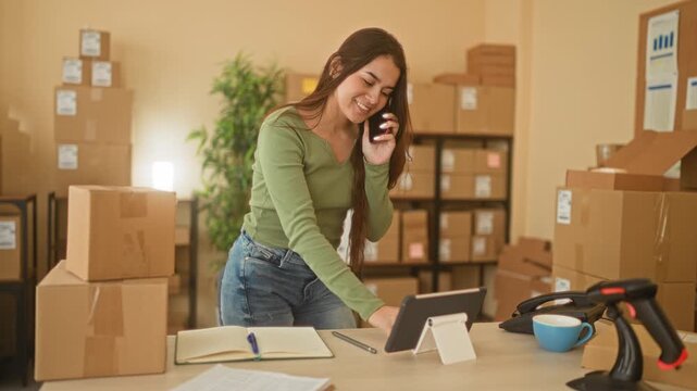 Woman talking on phone while tapping a tablet and organizing parcels at a packing desk in building; entrepreneurial focus.