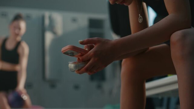 Close Up of Young Female Athlete Hands as Sits on Gym Bench. Dressed in Black Activewear, Girl Holds Pink Smartphone and Case For Wireless Bluetooth Earbuds. Interaction With Modern Technology.