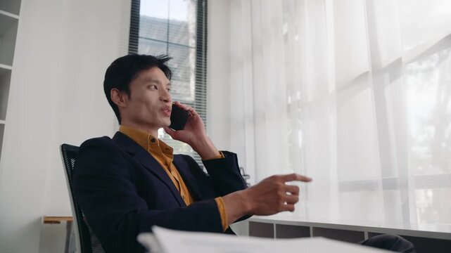 Confident young asian male manager in a suit speaking on a smartphone. He is sitting at his desk near a window in a modern office