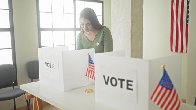 Woman smiles at a voting booth next to small american flags and an i voted sticker while standing inside a public building; pride civic duty.