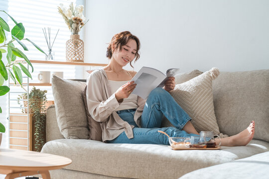 A stylish Asian woman sitting on the living room sofa, reading a magazine during teatime.