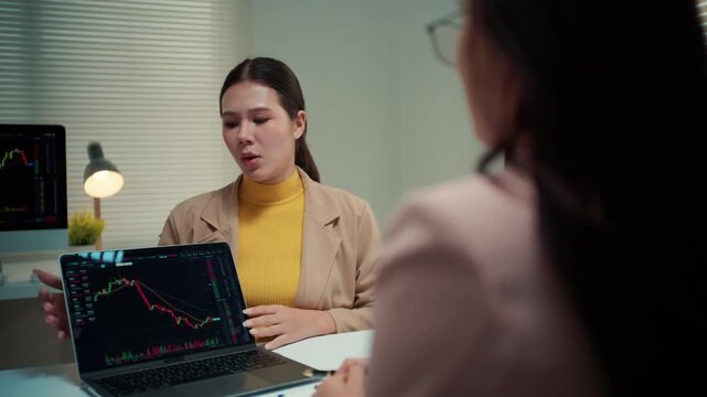 Asian businesswoman discussing investment strategy with a colleague, pointing at a bearish candlestick chart on a laptop screen in the office