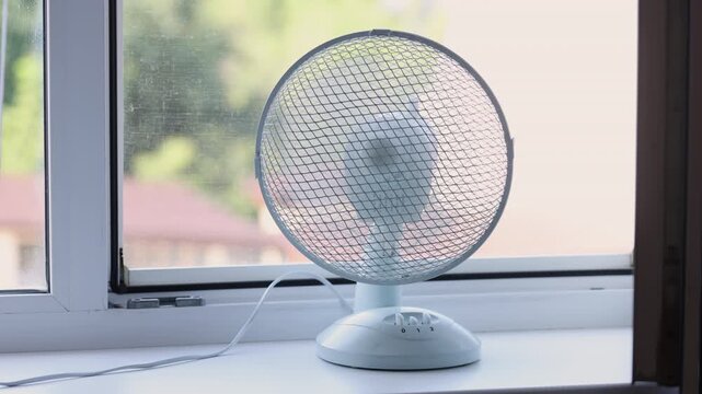 Electric fan blowing air on desk near window in sunny room. Household appliance cooling during extreme heat wave. Summer weather climate control and ventilation equipment concept.