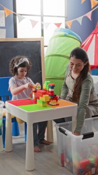 Woman stacks colorful blocks on a white table with a child, assembling a tower while seated in a playroom full of toys; nurturing patience.