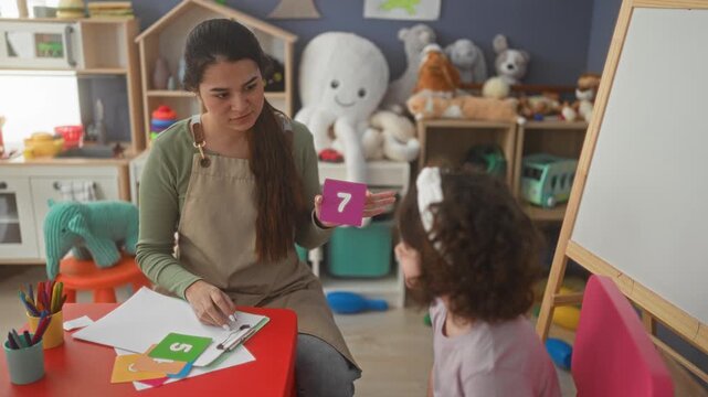 Woman teacher holds purple number 7 flashcard and points with hand to cards on table in a school building; early learning calm.