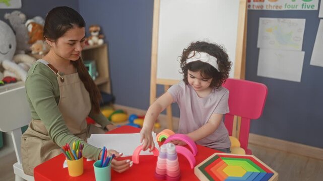 Teacher hands paper and smiles while girl stacks rainbow toy with her hands at red table in classroom building; playful learning.
