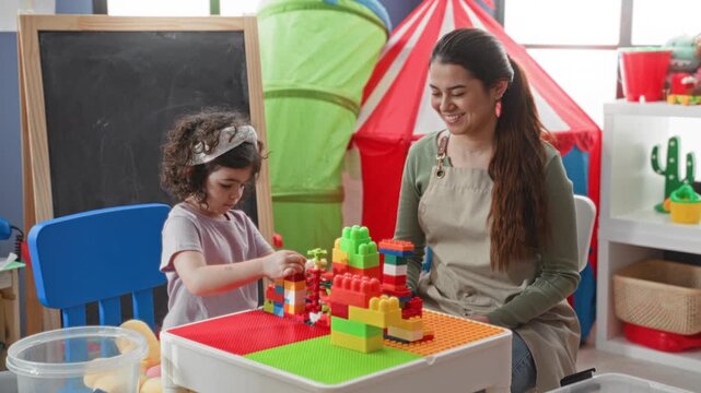 Woman sits with girl at low table using hands to stack colorful plastic building blocks beside chalkboard and play tent in preschool classroom; learning joy.