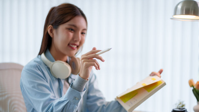 Young Asian student holding smartphone and notebook while studying with headphones in bright home workspace with copy space