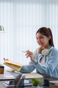 Young Asian female student smiling while reviewing colorful study materials with headphones at desk with copy space