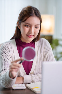 Young Asian student holding magnifying glass examining information on laptop during online education session at home desk