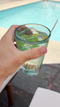 Point of view vertical video hand holding cold fresh cocktail by swimming pool. Enjoying summer vacation outdoors. Tourist in hotel resort holiday vibe. Vlogger content on social media travel fun reel