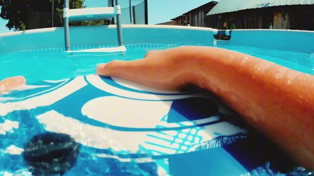 First-person perspective of a individual holding onto a blue and white floatie inside an above-ground pool during summer. Ideal for family recreation and leisure advertising.