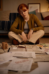 Fototapeta na wymiar Vertical shot of young adult Caucasian woman reviewing bills and notes while sitting on floor, managing household budget and financial stress. Ideal for debt, budgeting, personal finance marketing