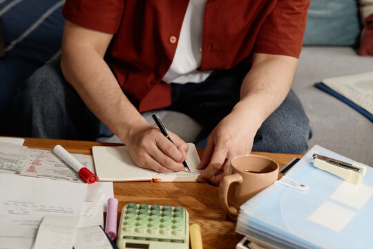 Close up of young adult man writing budget notes among bills, calculator, documents, managing household finances and planning expenses. Ideal for personal finance or debt content