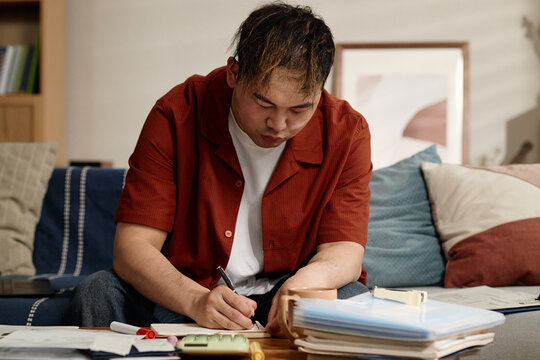 Asian young adult man writing notes and reviewing documents at home, managing paperwork and budgeting tasks. Useful for personal finance, debt planning, remote admin concepts