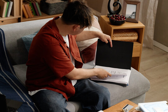 Young adult man reviewing paperwork on sofa, checking contract pages and organizing documents for home administration. Useful for insurance, finance, legal, budgeting themes