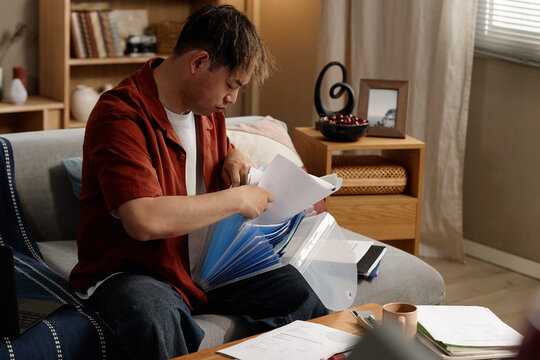 Young adult man sorting documents from expanding file folder while sitting on sofa, managing home paperwork and organization. Useful for tax filing, billing, admin, remote life