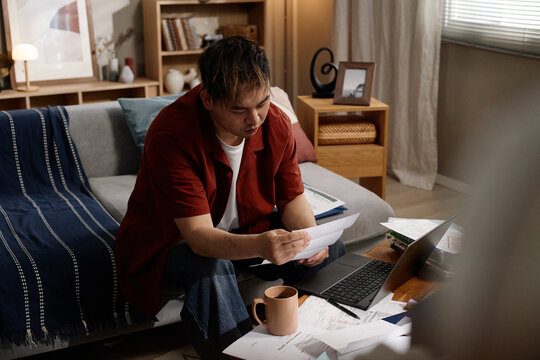 Young Asian man reviewing bills and paperwork beside laptop in living room, managing household finances and budgeting. Useful for debt, personal finance, remote admin themes