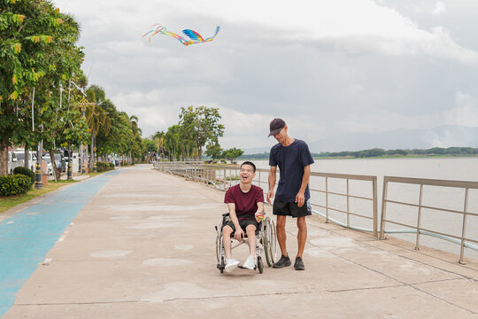 Happy young disabled man in wheelchair laughing with male caregiver while flying a colorful kite on a lakeside walkway, inclusive outdoor lifestyle, diversity and support concept.