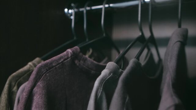 Close up man hand browsing through row assorted casual t shirts hanging on black hangers in dark wardrobe, carefully making selection for daily outfit. Man choosing clothes hanging on rack in closet