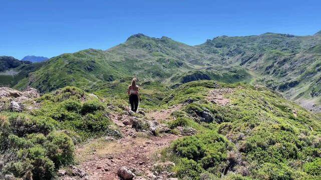 Woman hiker walking on a mountain trail surrounded by green hills under a clear blue sky