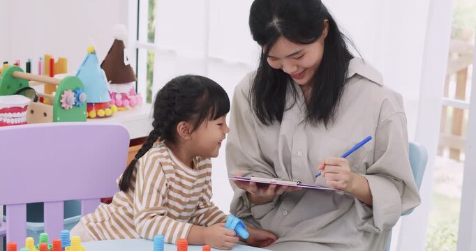 Asian mother or teacher holding a clipboard and taking notes while little girl playing with educational toys.