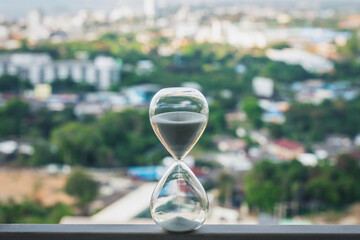 Fototapeta premium Hourglass standing on balcony railing with blurry cityscape background. Concept of time management, deadline countdown, urban lifestyle pace, and future planning.