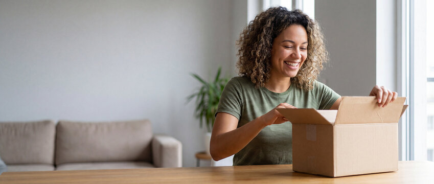Happy woman opening delivery box at home, sitting at a wooden dining table in a bright modern living room, showing joyful excitement for online shopping and e-commerce lifestyle concepts
