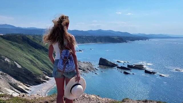 Woman tourist with backpack holding straw hat on cliff looking at scenic ocean coast in asturias spain