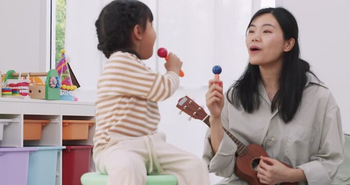 Happy Asian mother playing ukulele while her little daughter sings with toy microphone at home.
