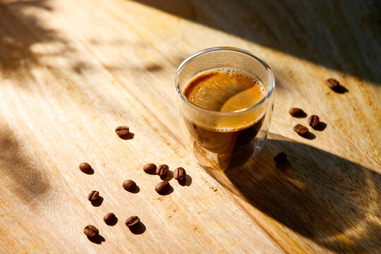 Fresh espresso coffee in a double wall glass cup illuminated by warm morning sunlight on a rustic wooden table.