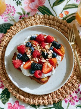 Overhead shot of French toast topped with fresh berries, whipped cream, and almonds on a decorative plate, surrounded by a floral tablecloth and woven placemat
