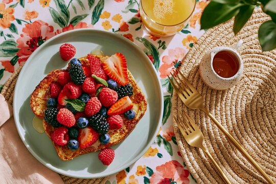 Overhead shot of a summer brunch table featuring French toast topped with fresh berries, a glass of juice, and decorative tableware on a floral tablecloth