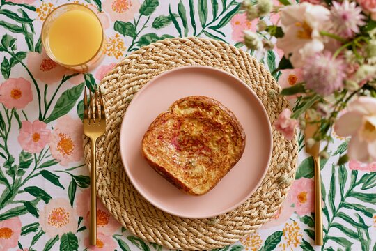 Overhead shot of a full breakfast table featuring golden French toast on a pink plate, surrounded by a woven placemat and a glass of orange juice