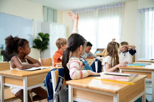 Bright Asian schoolgirl raising her hand to volunteer an answer during a classroom lesson. Confident elementary student raising her hand for participation in class with classmates.