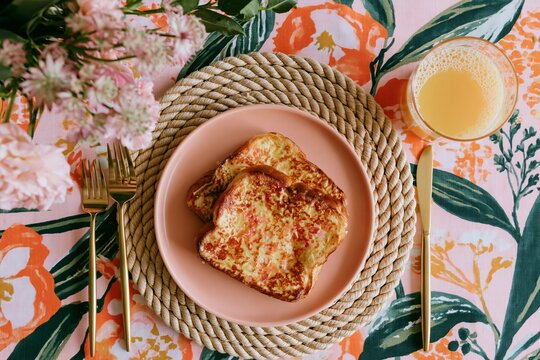 Overhead shot of a full breakfast table featuring French toast on a pink plate, surrounded by floral tablecloth, cutlery, and a glass of orange juice
