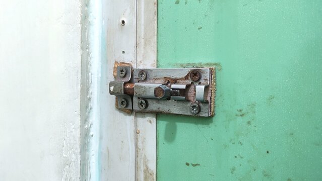 Close-up photograph of a metal door latch lock mounted on an old green door, showing security hardware with visible signs of rust, wear, and everyday use. Suitable for concepts of home security