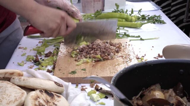 Close-up shot of a hand using a cleaver to chop green peppers on a wooden board, with pre-chopped roasted meat piled alongside